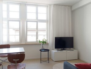 Een moderne woonkamer met een flatscreen televisie, een eettafel met stoelen, een klein bijzettafeltje met een vaas en een raam met gordijnen.