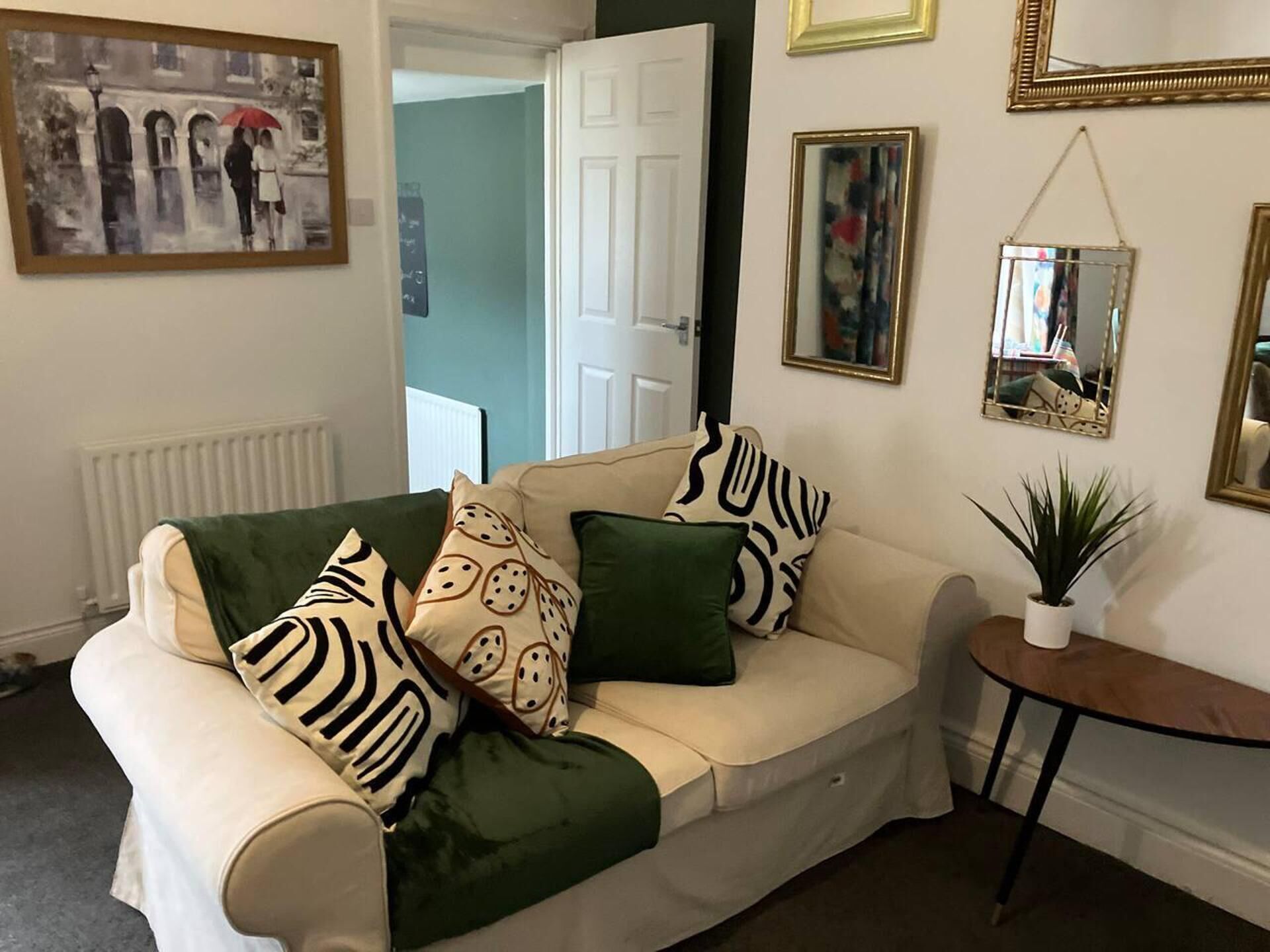 A living room with a beige sofa, green throw, and patterned pillows. There are wall-mounted mirrors and a framed painting. A small table with a plant is visible.