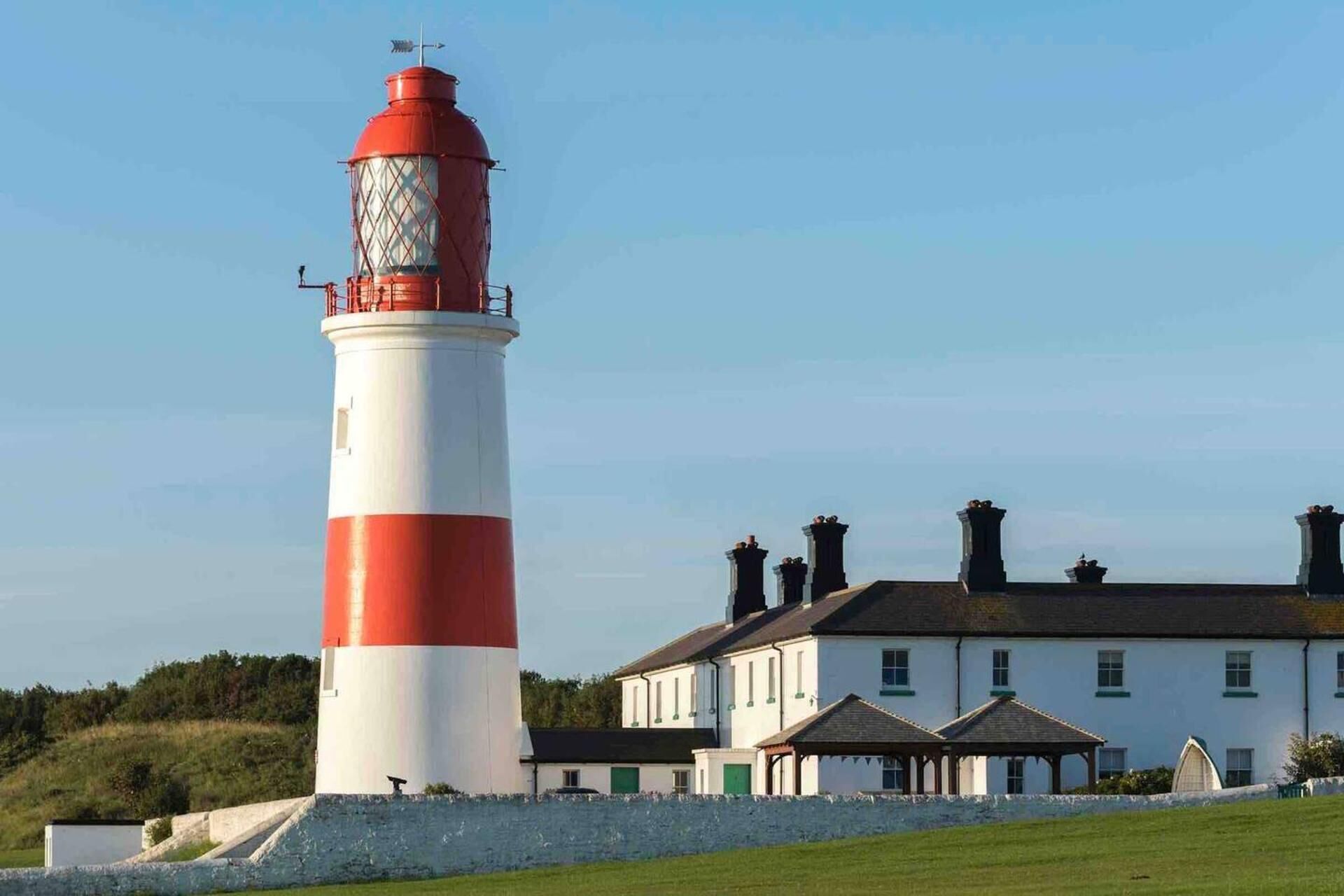A lighthouse with a red and white striped tower next to a white building with multiple chimneys and a green roof.