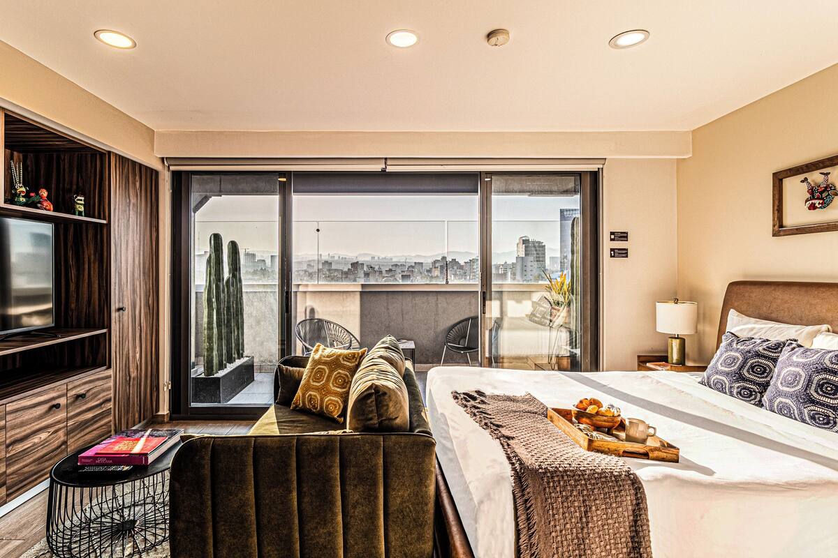 Quarto de hotel com cama, sofá, televisão, varanda com vista para a cidade e área de refeições com uma bandeja de comida.