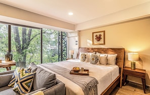 A hotel room with a large bed, a sofa, a nightstand, and a window with a view of trees.