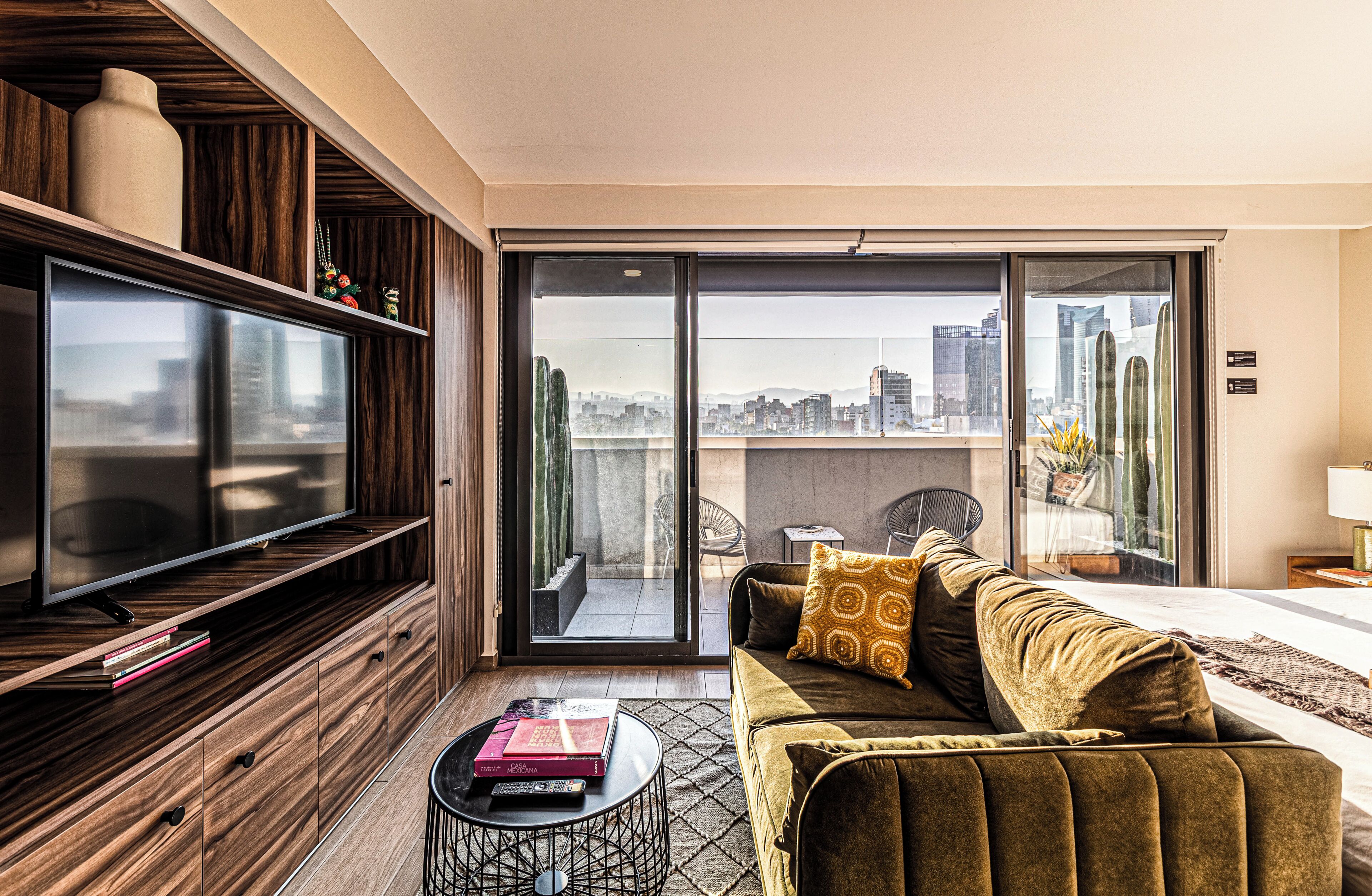A modern living room with a flat-screen TV, a sofa with patterned cushions, and a city view through large sliding glass doors.