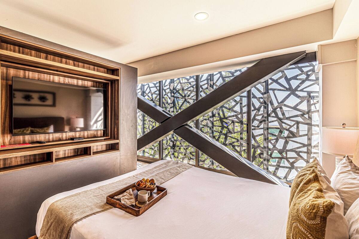 Quarto de hotel moderno com uma cama grande, uma TV fixada na parede e uma janela com tela metálica geométrica.
