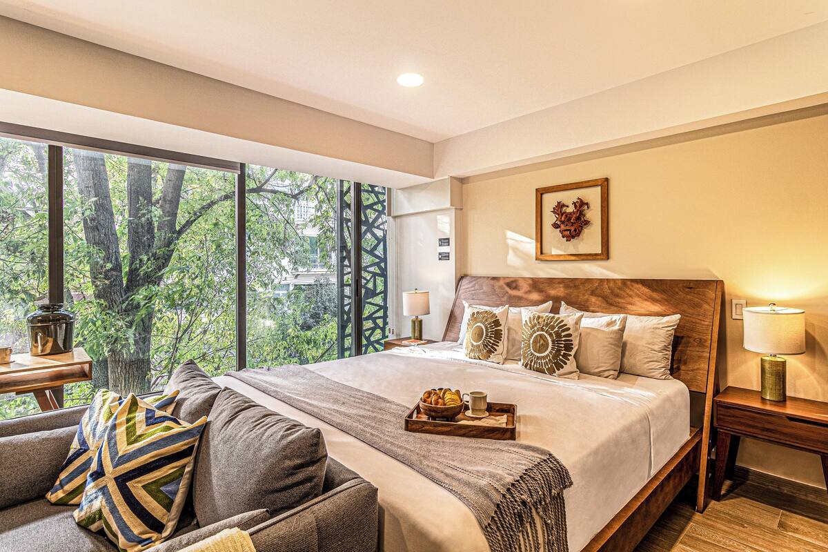 Quarto de hotel com uma cama grande, um sofá, uma mesinha de cabeceira e uma janela com vista para árvores.