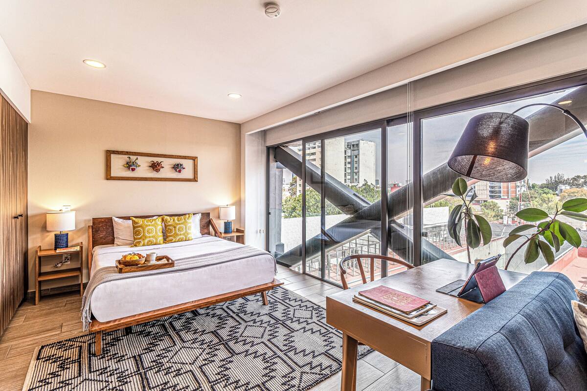 Quarto de hotel moderno com uma cama grande, uma escrivaninha com cadeira, uma janela com vista para edifícios e uma escada que leva a uma área externa.