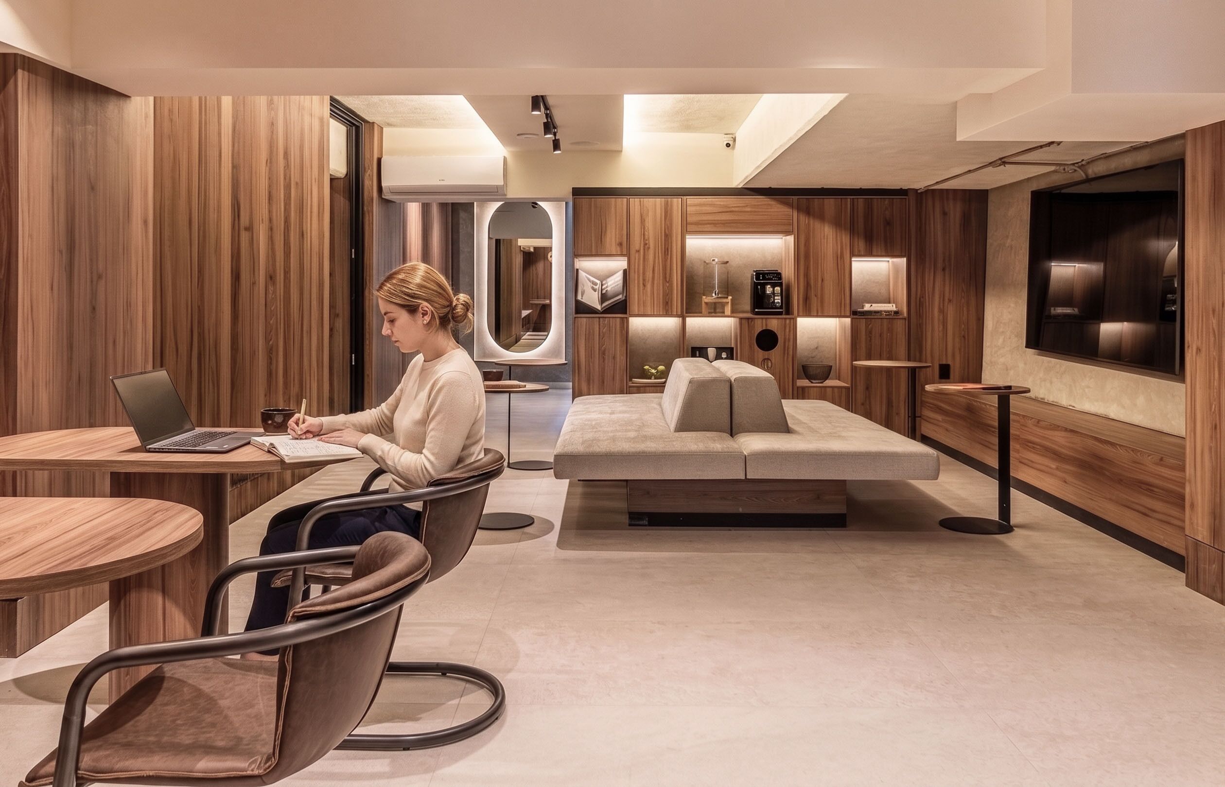 A modern hotel room with a wooden panel wall, a sofa, a desk with a laptop, and a TV mounted on the wall.