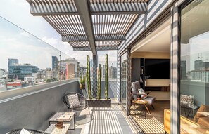A modern balcony with a city view, featuring a seating area with chairs and a table, potted plants, and a glass door leading to an interior space. - Kukun Tonalá 30 Roma (Mexico City)
