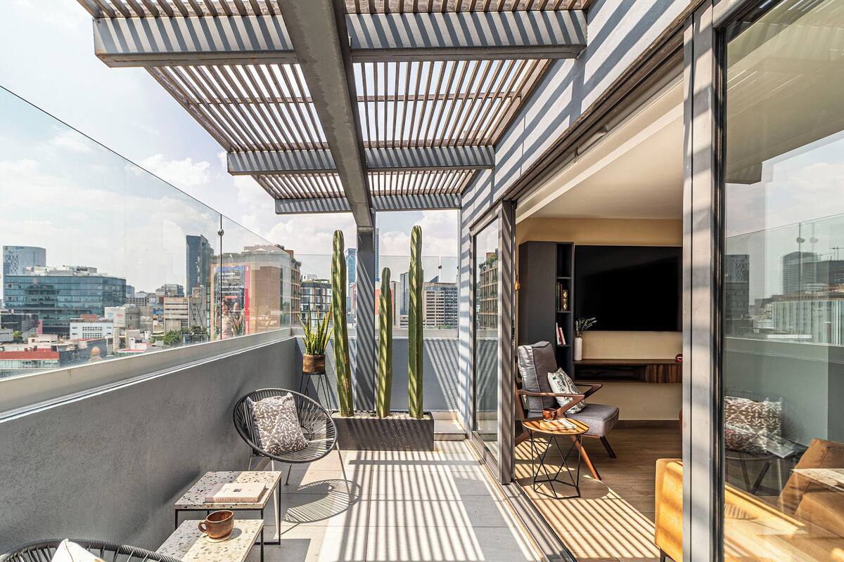 Varanda moderna com vista para a cidade, com área de estar, cadeiras, mesa, plantas em vasos e porta de vidro que dá para um espaço interior.