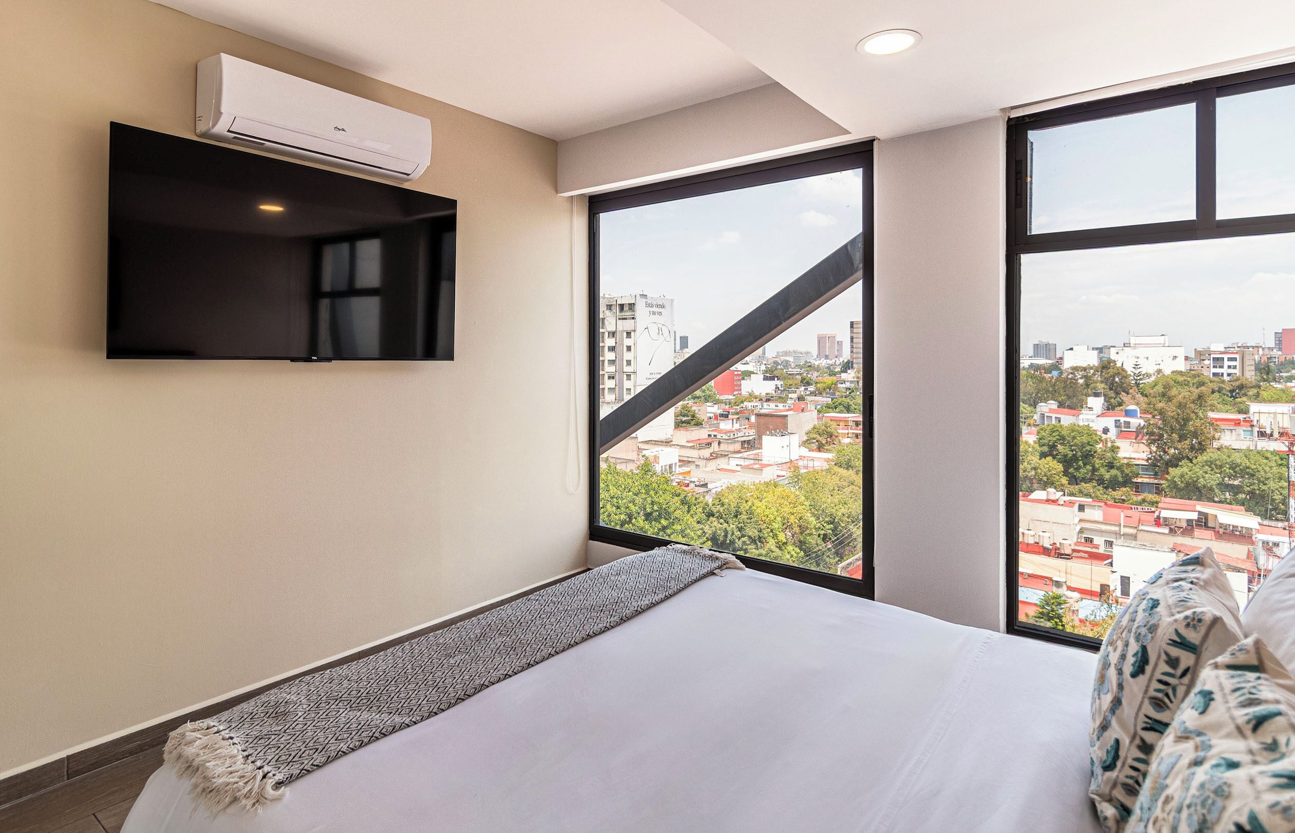 Een slaapkamer met een bed, een wandairconditioner, een televisie en een groot raam met uitzicht op de stad.