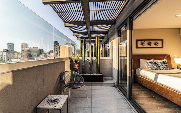 A balcony with a city view, a bed with patterned pillows, a potted plant, a chair, and a table with a book.