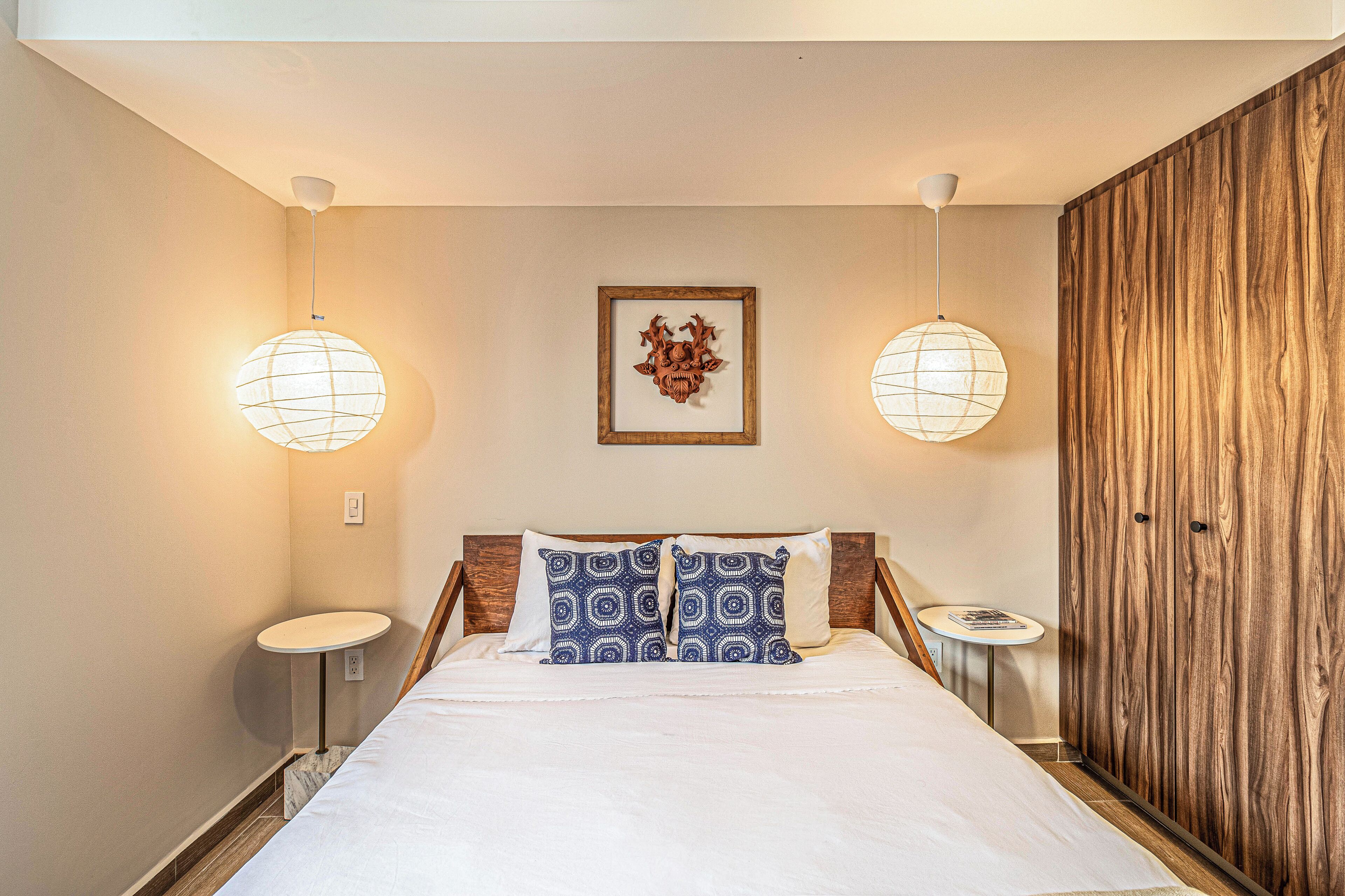 Un dormitorio con una cama, dos mesitas de noche, una lámpara de pared, una obra de arte enmarcada y un armario de madera.
