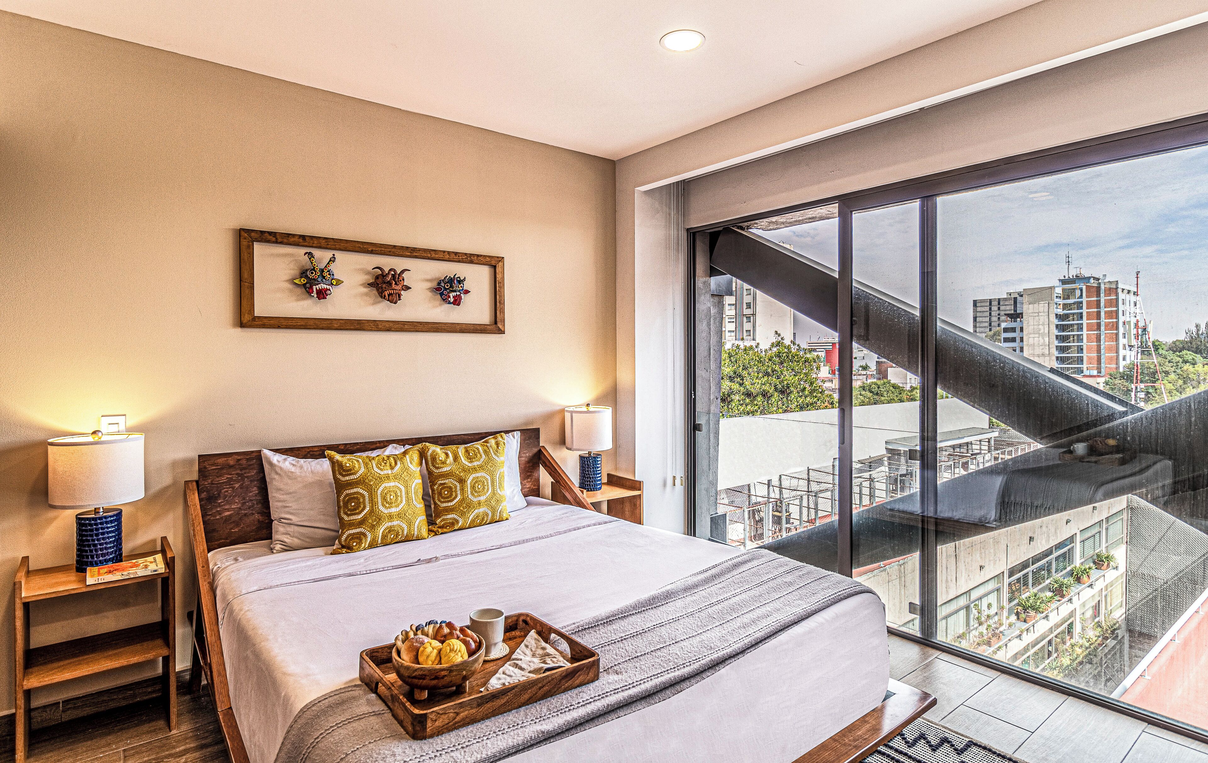 A hotel room with a large bed, two bedside tables, a tray with pastries, a framed artwork above the bed, and a city view through a large window.