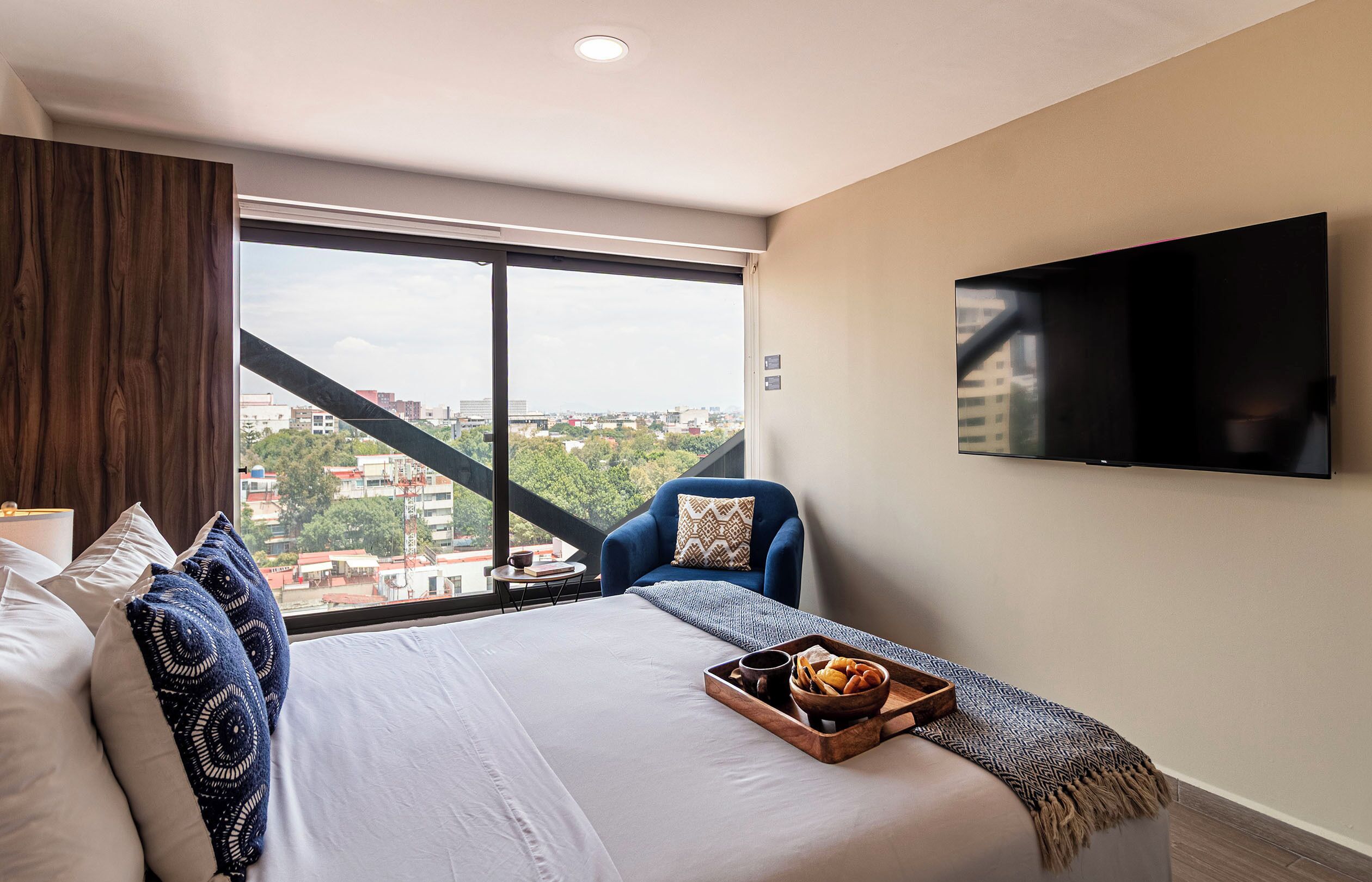 Ein Hotelzimmer mit einem großen Bett, einem an der Wand montierten Fernseher, Blick auf die Stadt durch ein großes Fenster und einem kleinen Tisch mit einer Speisewagenaufsatz.