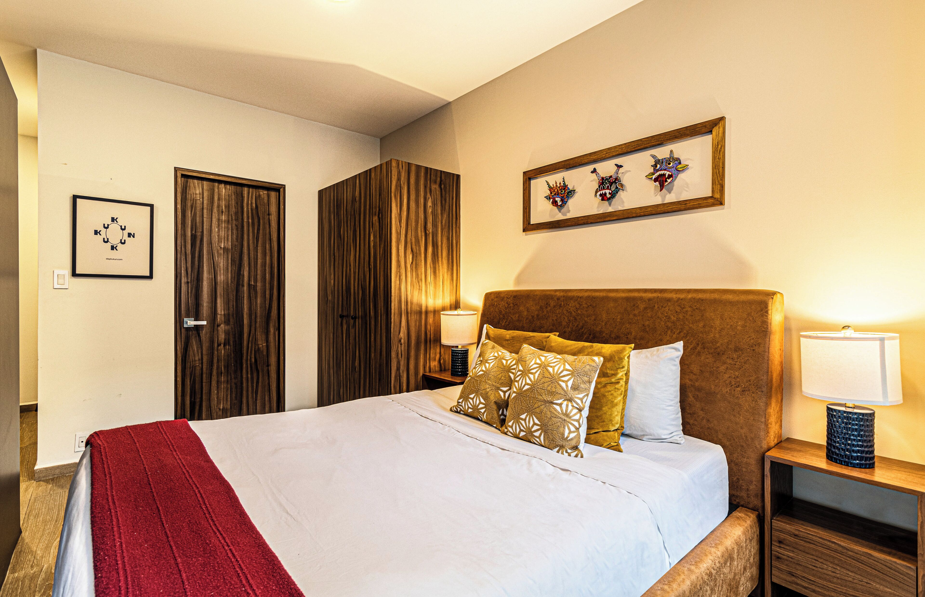Quarto de hotel com uma cama, um guarda-roupa de madeira, uma mesa de cabeceira com abajur e um quadro na parede.