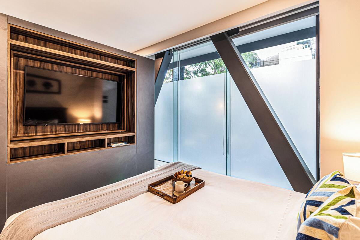 Quarto de hotel com cama grande, móvel de TV de madeira, porta de vidro que dá para o exterior e bandeja com xícara e doces sobre a cama.