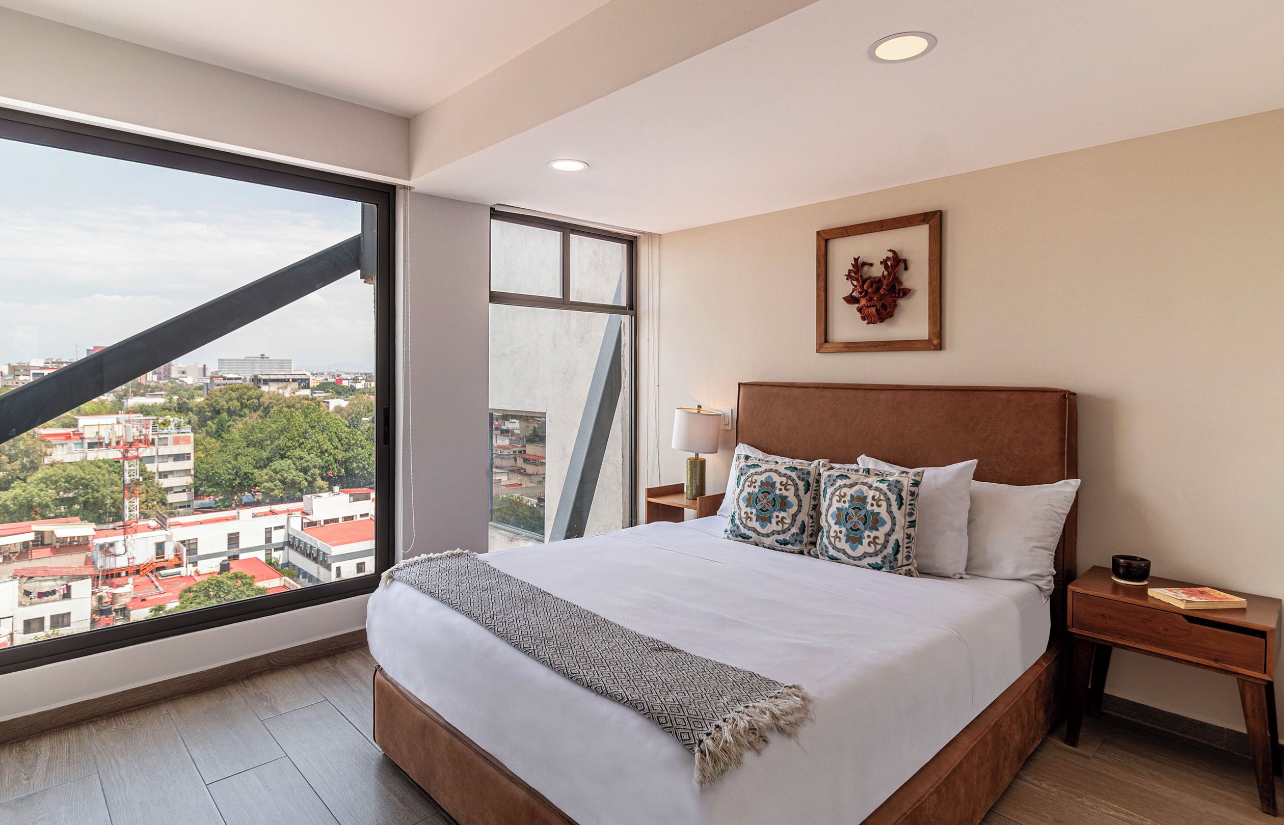 A hotel room with a large bed, a nightstand, a bedside lamp, a framed picture on the wall, and a view of the city through a large window.