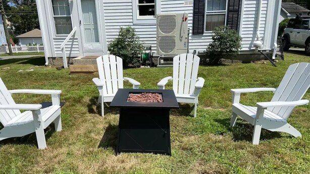 Un patio trasero con un pozo de fuego rodeado de sillas Adirondack blancas.