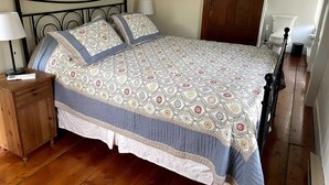 Una cama bien hecha con una colcha estampada y almohadas, una mesita de noche de madera con una lámpara y piso de madera.