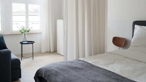 Une chambre d’hôtel moderne, équipée d’un lit, d’une petite table ornée d’un vase de fleurs, d’une fenêtre avec des rideaux et d’un haut-parleur fixé au mur.