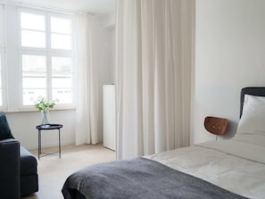Een moderne hotelkamer met een bed, een klein tafeltje met een bloevas, een raam met gordijnen en een wandluidspreker.