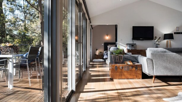 A modern living room with a large glass door leading to a deck, a grey sofa, a wooden coffee table, and a flat-screen TV mounted on the wall.