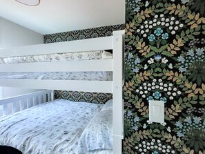 A bunk bed with patterned bedding, a white wall with a floral wallpaper, and a light switch.