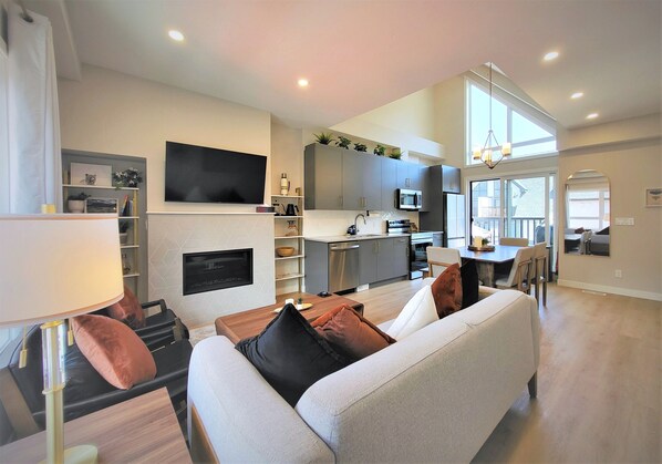 A modern living room with a sofa, a coffee table, and a TV. There is a kitchen area with cabinets and a microwave.