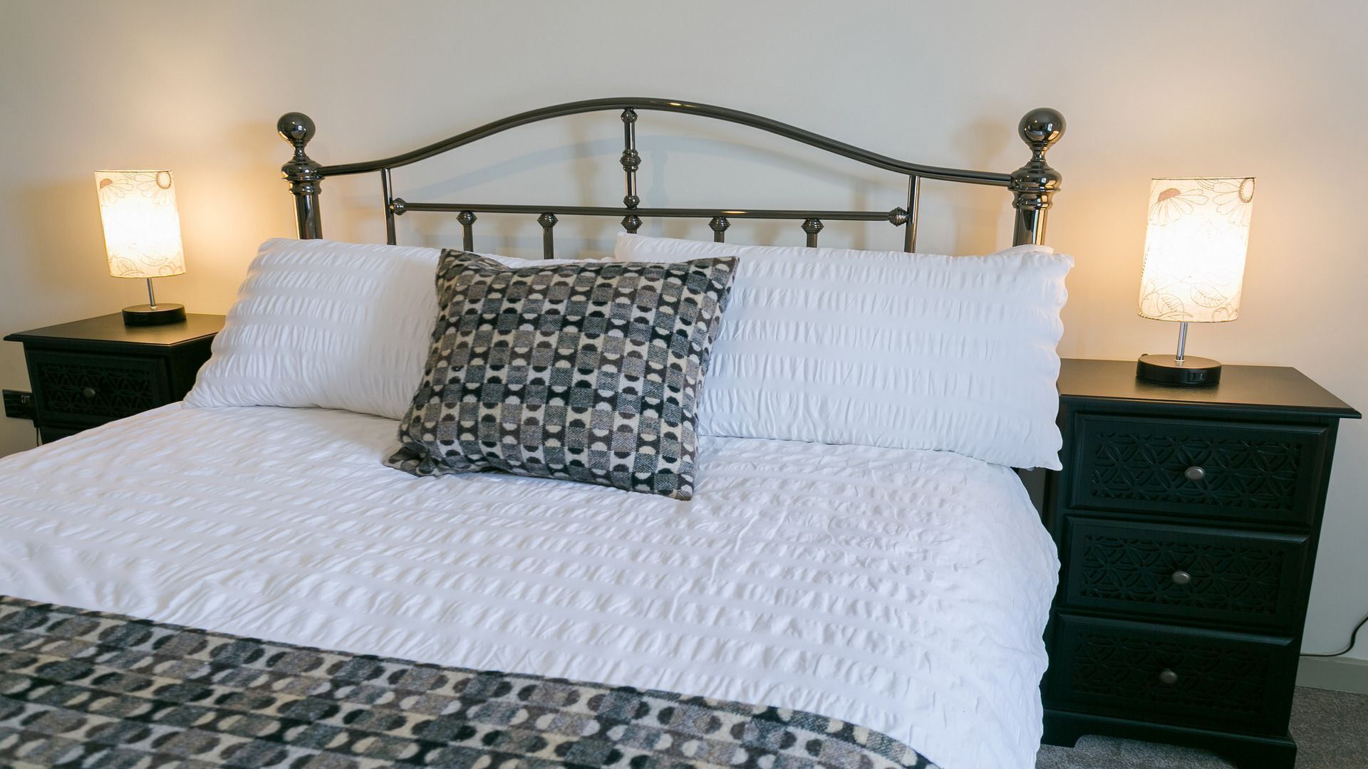 Un lit bien fait, avec une literie blanche et un coussin à motifs, flanqué de deux tables de chevet sur lesquelles sont posées des lampes.