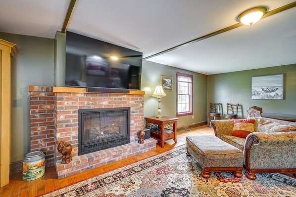 A living room with a brick fireplace, a television mounted above it, a patterned rug, and a sofa with an ottoman. - Buskirk Vacation Rental w/ Deck & Putting Green! (Buskirk)