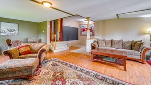 Een ruime woonkamer met een houten vloer, een grote bloemenbank, een passende fauteuil en een bijzettafeltje. Een trap met rode leuning leidt naar een ander niveau.