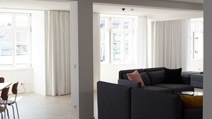 Un salon moderne avec un canapé gris, une table à manger avec des chaises et de grandes fenêtres ornées de rideaux blancs.