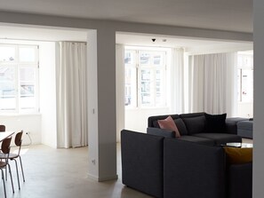 Een moderne woonkamer met een grijze bank, een eettafel met stoelen en grote ramen met witte gordijnen.