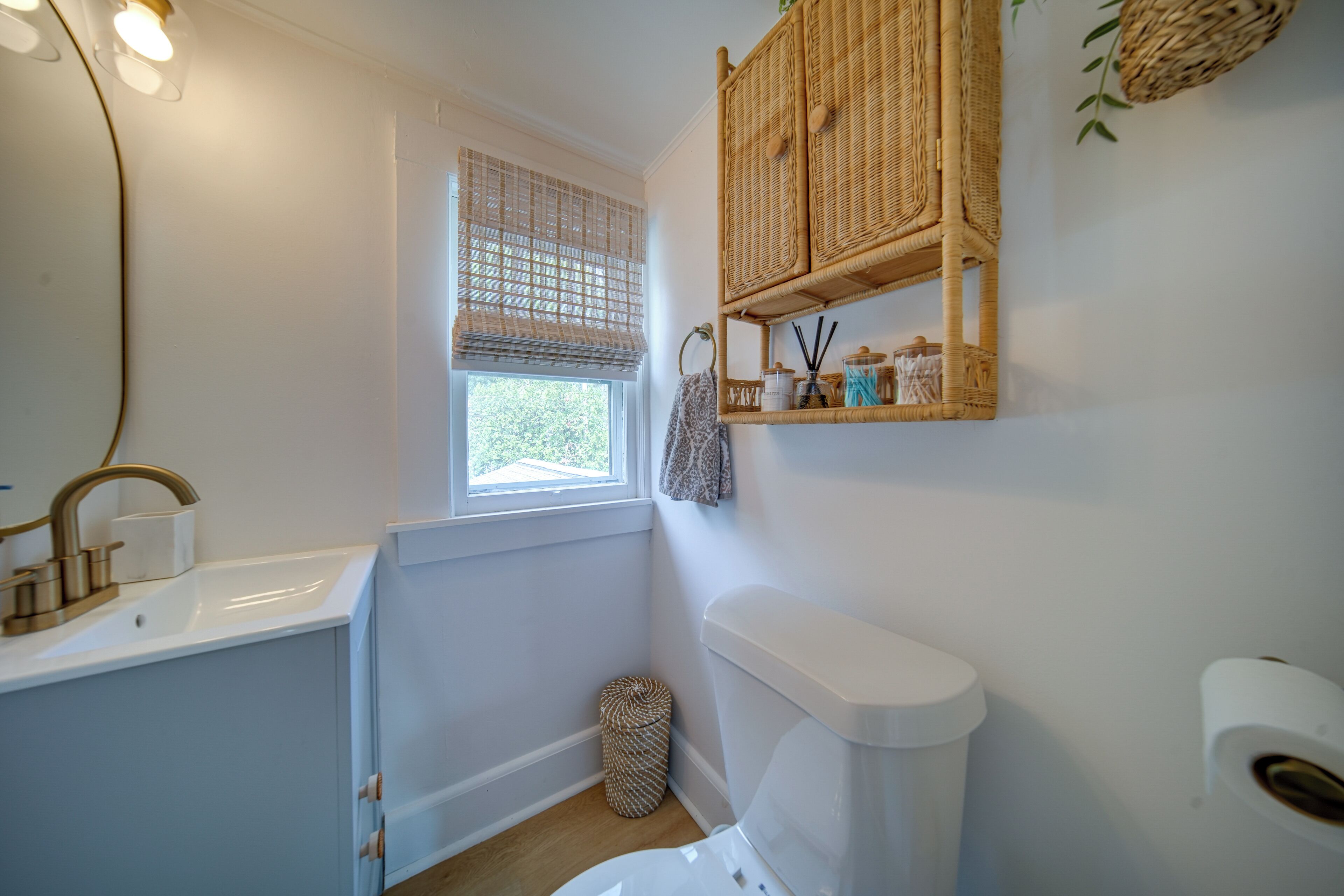 A bathroom with a white sink, a toilet, a window with a blind, and wicker baskets on the wall.