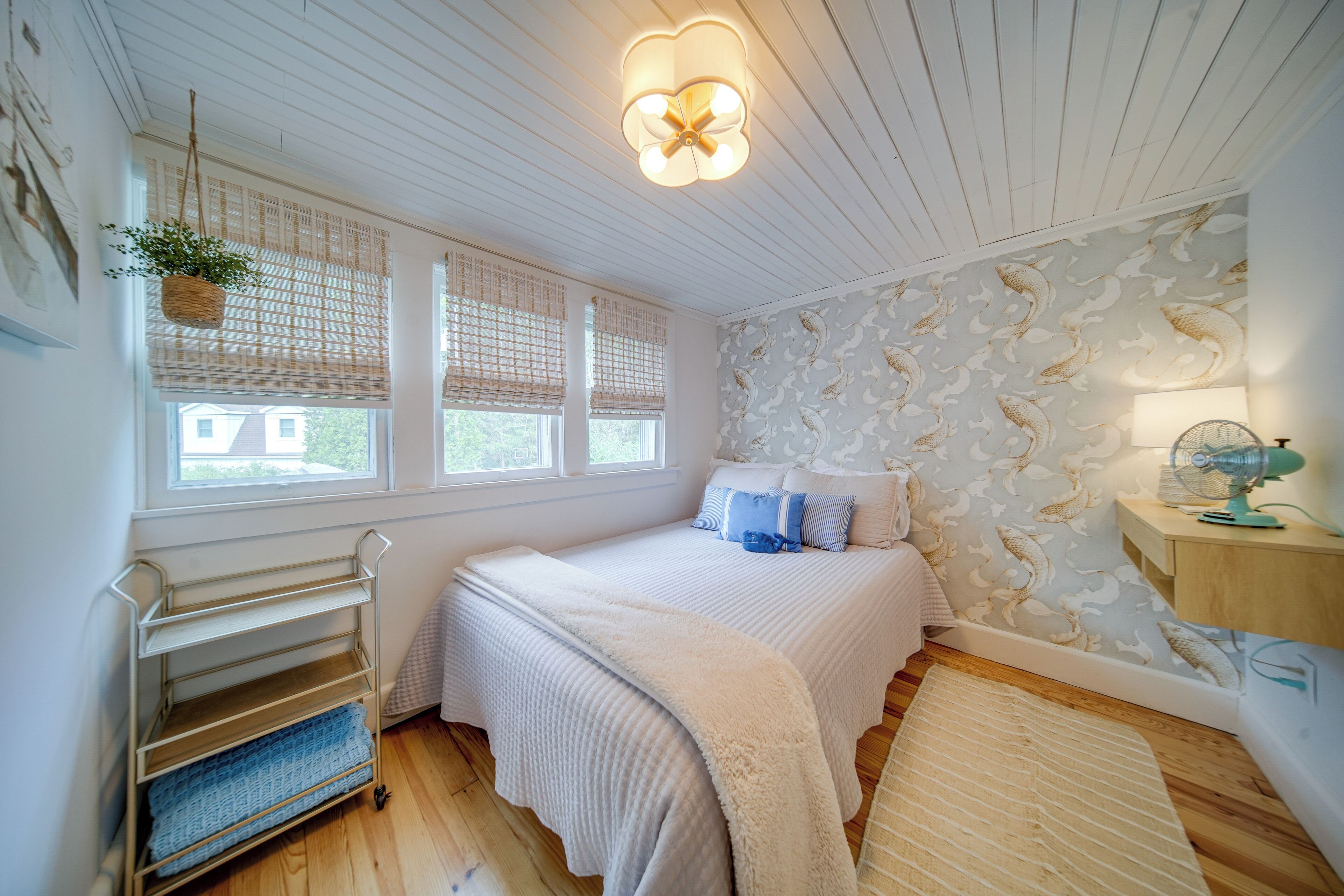 A bedroom with a bed, a bedside table, a lamp, a fan, a window with blinds, a hanging plant, and a wall with a fish pattern.