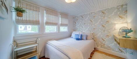 A bedroom with a bed, a bedside table, a lamp, a fan, a window with blinds, a hanging plant, and a wall with a fish pattern.