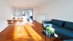 Un grand salon avec un canapé bleu, une table à manger avec des chaises, et une cuisine en arrière-plan.