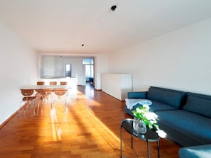 Een ruime woonkamer met een blauwe bank, een eettafel met stoelen, en een keuken op de achtergrond.
