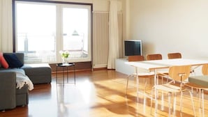 Un salon moderne comprenant un canapé, une petite table avec un vase, une table à manger avec des chaises, un téléviseur et une grande fenêtre avec des stores.