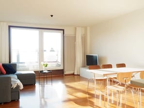 Een moderne woonkamer met een bank, een klein tafeltje met een vaas, een eettafel met stoelen, een televisie en een groot raam met jaloezieën.
