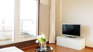 Un salon moderne avec une grande fenêtre, un téléviseur à écran plat posé sur un meuble blanc, une table ronde noire agrémentée d’un vase de fleurs et un plancher en bois.