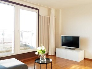 Een moderne woonkamer met een groot raam, een flatscreen televisie op een witte kast, een ronde zwarte tafel met een bloevas, en een houten vloer.