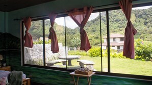 Une pièce dotée d’une grande fenêtre, de rideaux, d’un lit, d’une petite table et offrant une vue sur un jardin et des montagnes.