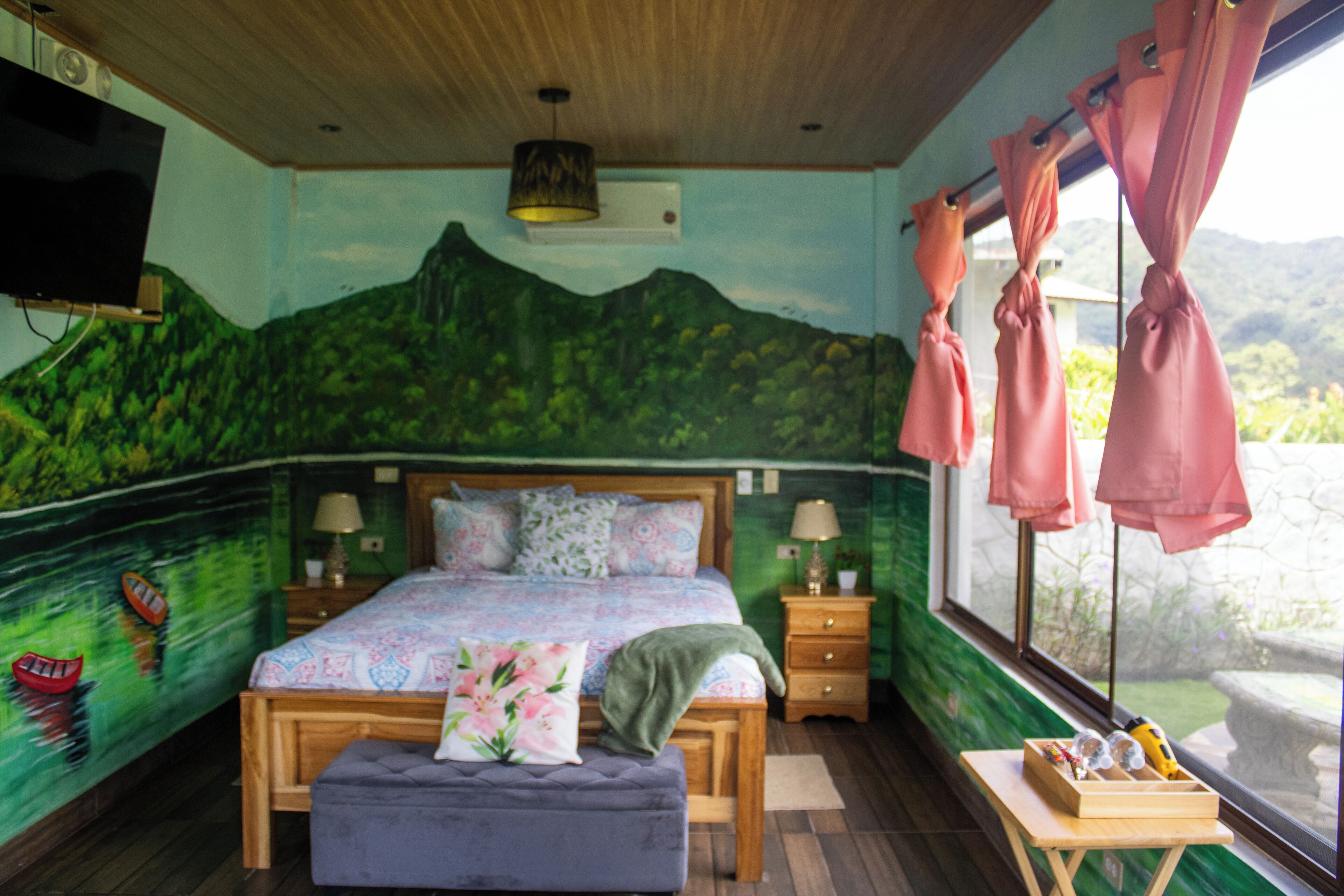 Een slaapkamer met een bed, nachtkastjes, een televisie, een raam met gordijnen en uitzicht op groen.