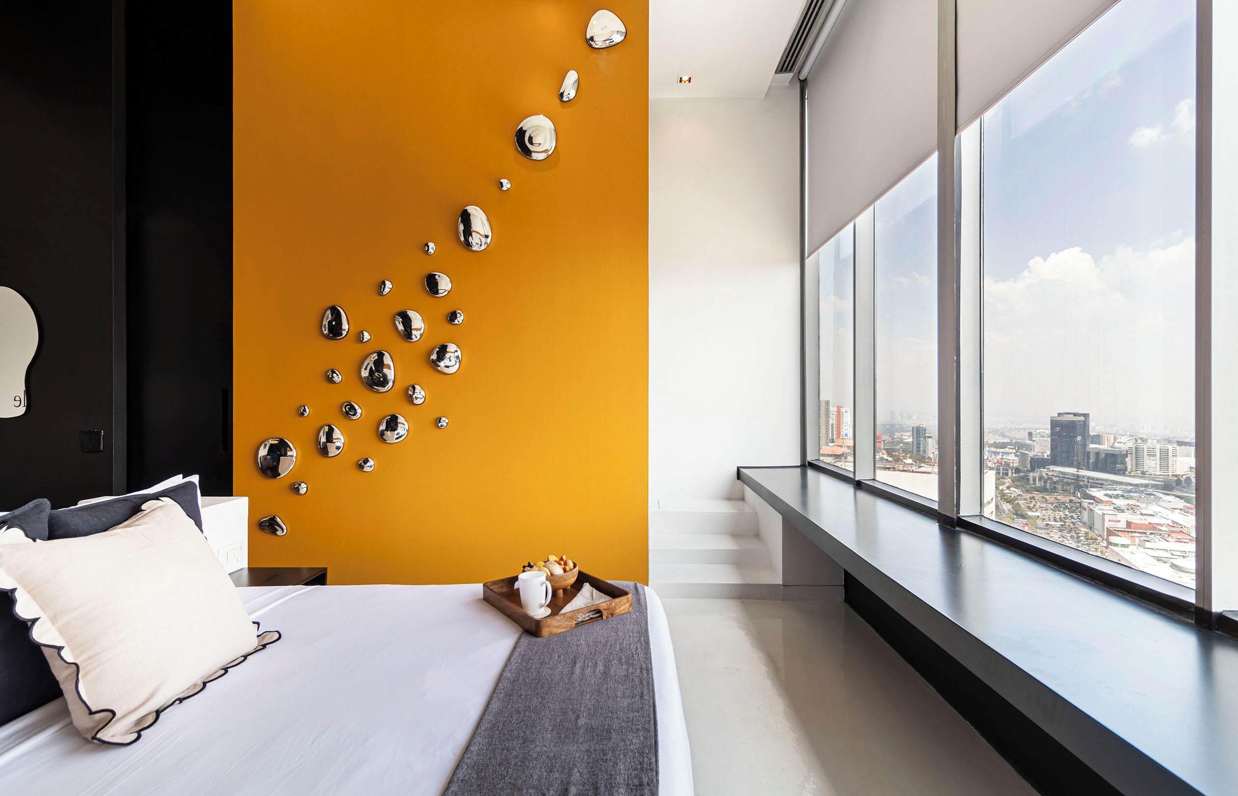 A modern hotel room with a large bed, a city view, and a unique wall decoration of reflective spheres.