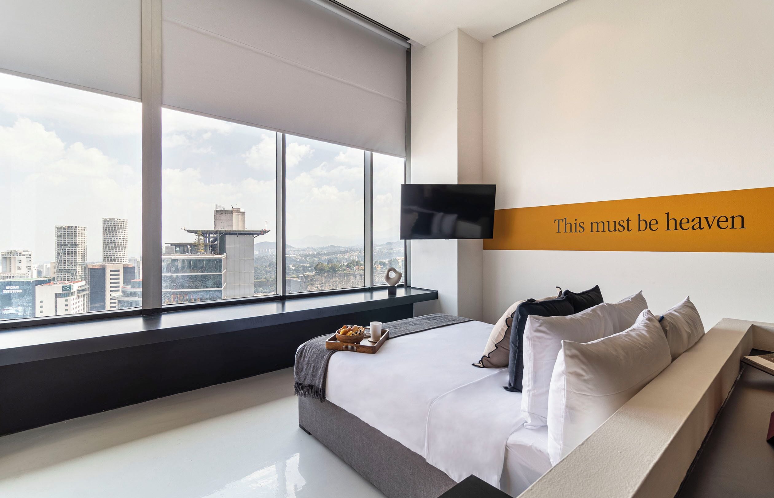 A modern hotel room with a large bed, a flat-screen TV mounted on the wall, and a city view through large windows.