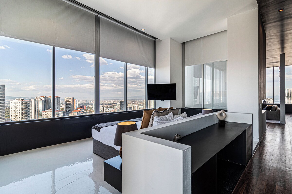 A modern hotel room with a large window offering a city view, a comfortable seating area, and a flat-screen TV mounted on the wall.