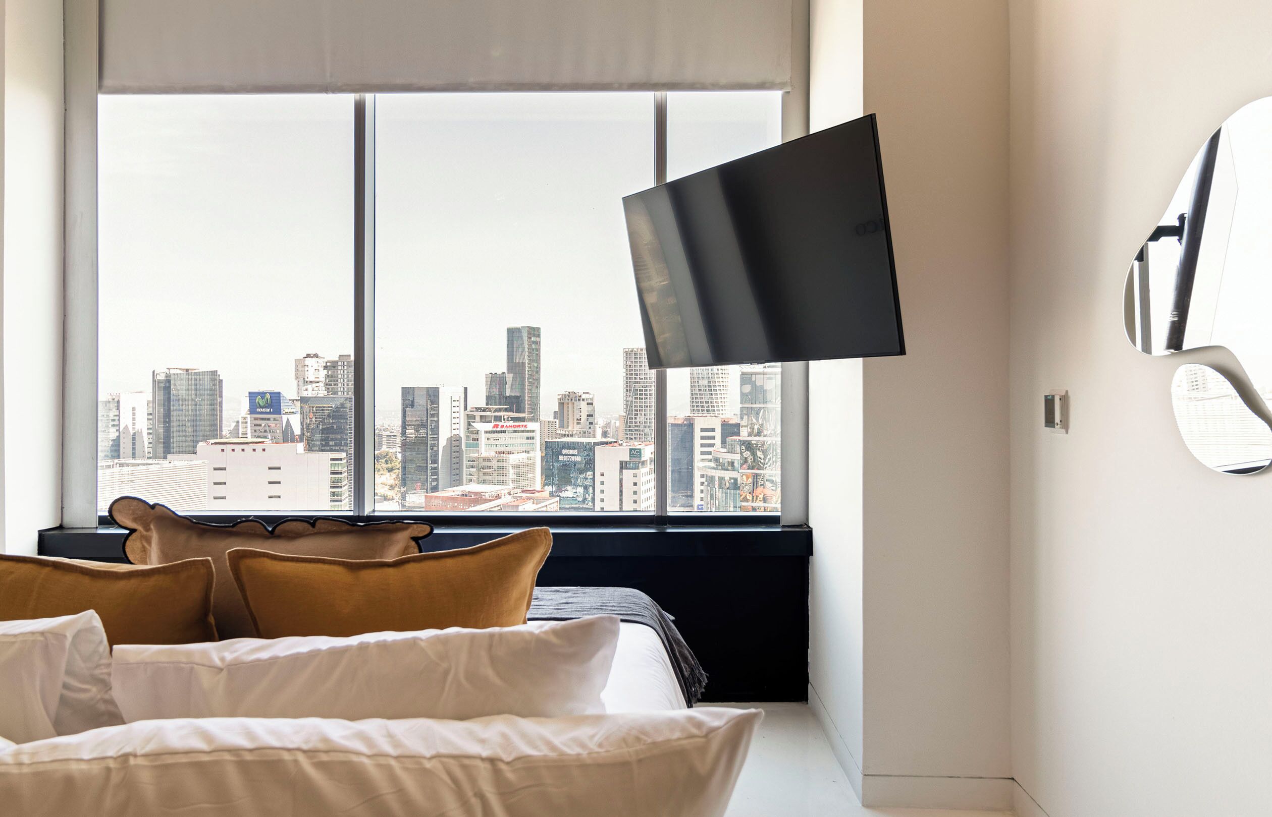 Una habitación de hotel moderna con un ventanal grande, una cama con ropa blanca y un televisor de pantalla plana montado en la pared.