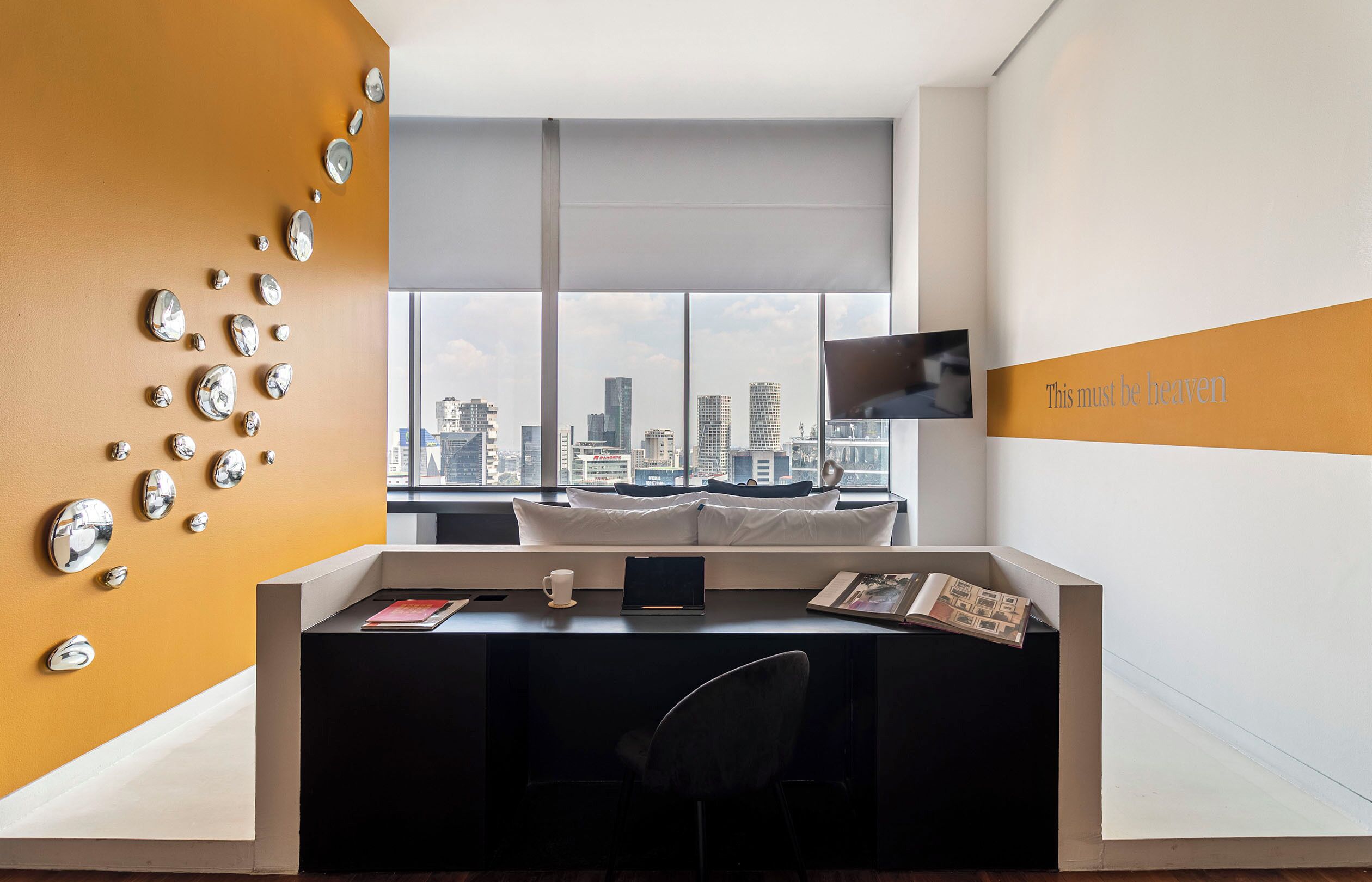 A modern office space with a city view, a desk with a laptop, a chair, and a wall-mounted TV.