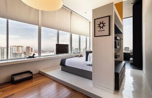 Habitación de hotel moderna con un ventanal grande, una cama, un televisor y vista a la ciudad.
