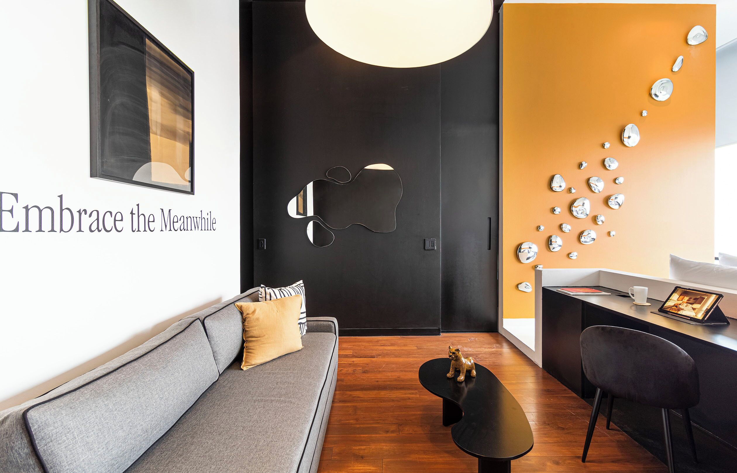 Een moderne hotelkamer met een grijze bank, een zwarte salontafel, een bureau met een laptop en een wand met een spiegelbeeld kunstwerk.